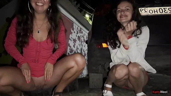 Two girlfriends pee together near a car in a public parking lot